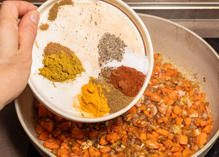Adding spices to a skillet with cooked onion and carrots.